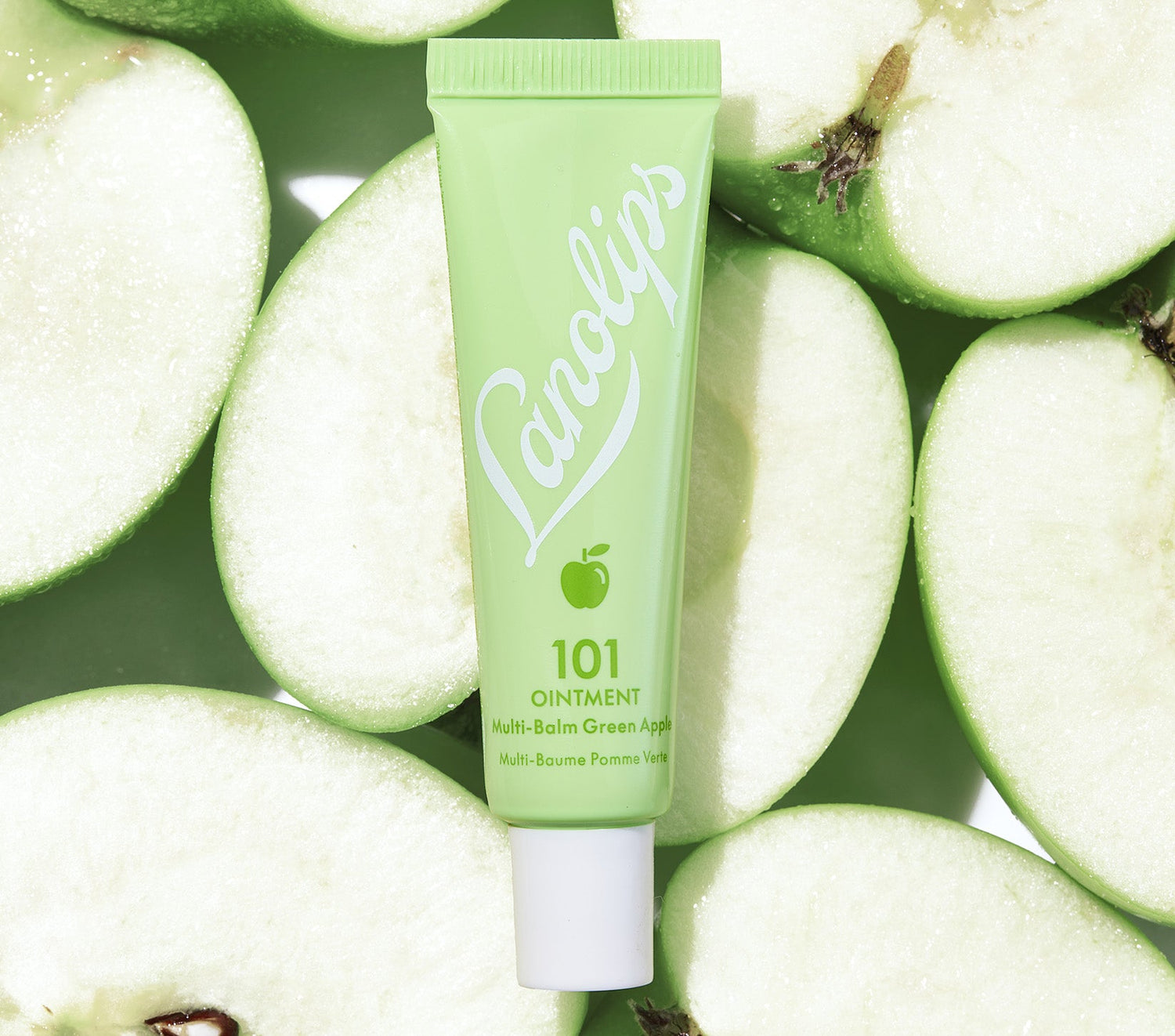 A tube of Lanolips 101 Ointment in the limited-edition Green Apple flavour is placed on a background of freshly sliced Granny Smith apples. The tube is a soft green shade with white branding and a small apple icon, reflecting the crisp and refreshing theme of the product.  This nourishing ointment contains Aussie lanolin, vitamin e and green apple extract, which mimics the skin’s natural oils to provide intense hydration, leave lips feeling nourished.